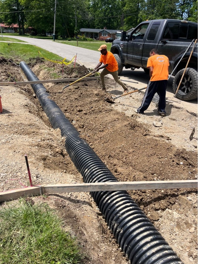 Concrete workers in orange shirts working around an excavated drainage area with a large pipe